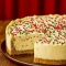 Close-up of a slice of sugar cookie cheesecake showing a golden cookie crust, creamy white filling studded with cookie-dough bits, glossy white-chocolate topping, and festive sprinkles.
