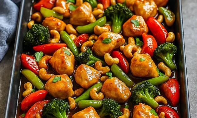 Sheet pan of bite-sized glazed chicken, roasted bell peppers, red onion and toasted cashews, served with a side of steamed rice.