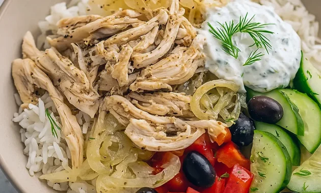 Shredded lemon-oregano slow cooker chicken piled on a bowl of rice with a side of tzatziki, sprinkled with parsley and lemon wedges.
