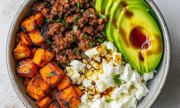 Overhead shot of a bowl with seasoned ground beef, roasted sweet potato cubes, sliced avocado, dollops of cottage cheese, and a honey drizzle, garnished with cilantro, Viral Hot Honey Ground Beef Bowl.