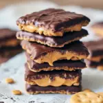 Stack of graham-cracker sandwiches filled with peanut butter, half-dipped in chocolate, on parchment with a sprinkle of chopped peanuts and flaky sea salt.
