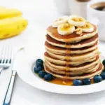 Golden stack of banana cottage cheese pancakes on a white plate, topped with banana slices, fresh berries and a light drizzle of maple syrup, with a fork and linen napkin beside the plate.