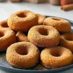 Close-up of cinnamon-sugar coated pumpkin donuts made with almond flour, stacked on a rustic plate with a sprinkle of cinnamon nearby.