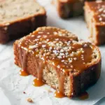 Sliced loaf of banana bread with visible caramel ribbons, drizzled salted caramel on top and a sprinkle of sea salt, on a wooden board.