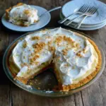 Slice of bourbon banana cream pie on a white plate, showing layers of golden cookie crust, banana slices, creamy custard, and a cloud of whipped cream with cookie crumb garnish, Dinner Desserts.