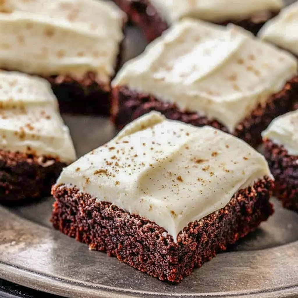 A platter of square gingerbread brownies frosted with creamy eggnog cream-cheese icing, dusted with cinnamon and arranged on a festive holiday napkin.