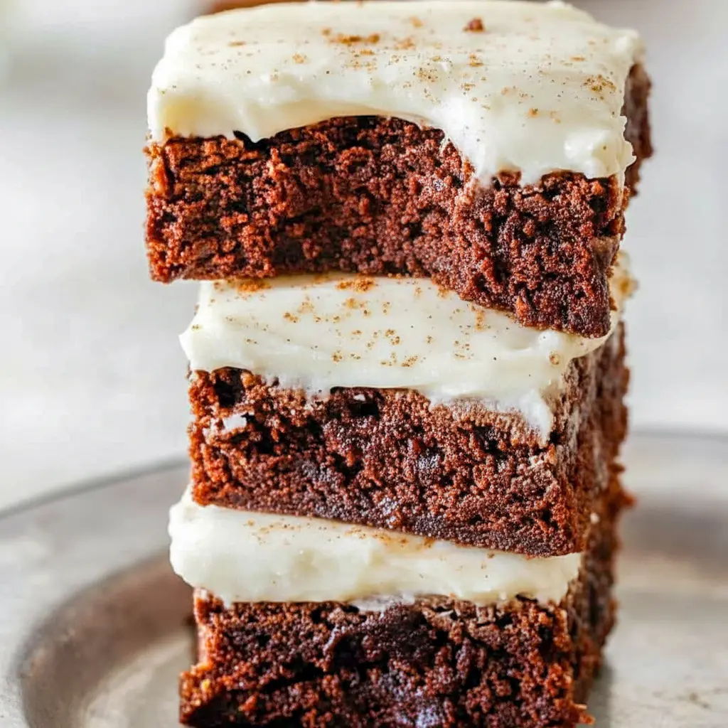 A platter of square gingerbread brownies frosted with creamy eggnog cream-cheese icing, dusted with cinnamon and arranged on a festive holiday napkin.