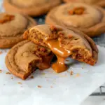 Warm, cracked Biscoff cookie showing a gooey molten cookie-butter center on parchment paper, golden edges and crumbs visible, Biscoff Lava Cookie.