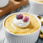 Three ramekins of creamy eggnog pudding topped with whipped cream, grated nutmeg, and sugared cranberries on a wooden tray, with a gold spoon nearby, Holiday Eggnog Pudding.
