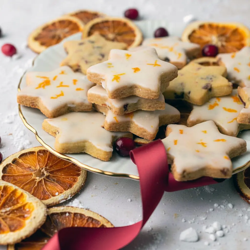 Overhead shot of star-shaped cranberry orange shortbread cookies glossed with orange glaze and sprinkled with zest, arranged on a wooden board with a festive ribbon nearby, Orange Christmas Recipes.