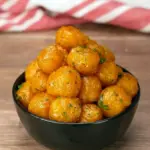 Close-up of golden garlic potato pops in a white bowl, glistening with melted butter and sprinkled with chopped parsley, ready to eat, Crispy Potato Snack Ideas.