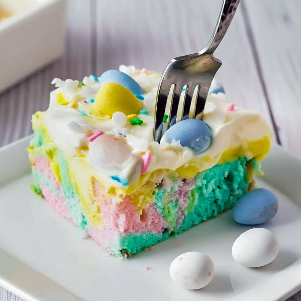 Close-up of a slice of pastel marbled poke cake with visible pudding-filled holes, topped with whipped cream, shredded coconut and mini chocolate eggs — a lovely option for Easter Sunday Dessert Ideas, a fresh Easter Spring Desserts pick, and an easy choice in Easy Desserts Easter, Fun And Easy Easter Desserts.