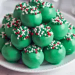 Close-up of glossy green truffles coated in candy melts with festive sprinkles; one truffle is cut open to reveal a creamy chocolate-cookie center, Christmas Truffles.