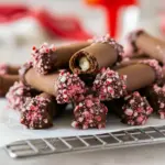 Close-up of a platter of rolled chocolate cookies filled with peppermint cream and sprinkled with crushed candy cane, styled with Dark Chocolate Peppermint Crunch Cookies, tiny decorative Chocolate Peppermint Spoons, and crumb-covered Chocolate Peppermint Crumble Cookies, Chocolate Peppermint Roll.