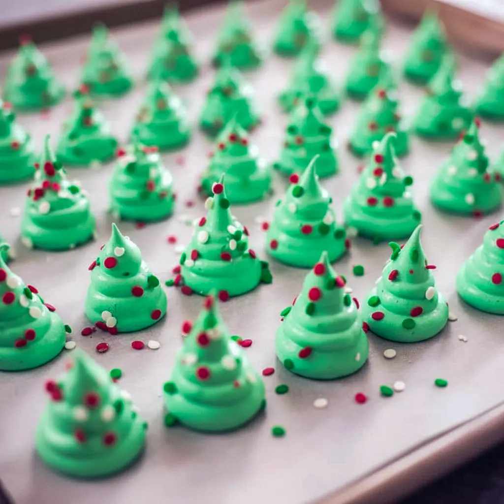Close-up of piped green meringue Christmas tree cookies on parchment, sprinkled with nonpareils and a few pretzel trunks visible, cooling on a sheet pan, Meringue Christmas Trees.