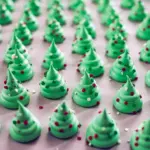 Close-up of piped green meringue Christmas tree cookies on parchment, sprinkled with nonpareils and a few pretzel trunks visible, cooling on a sheet pan, Meringue Christmas Trees.