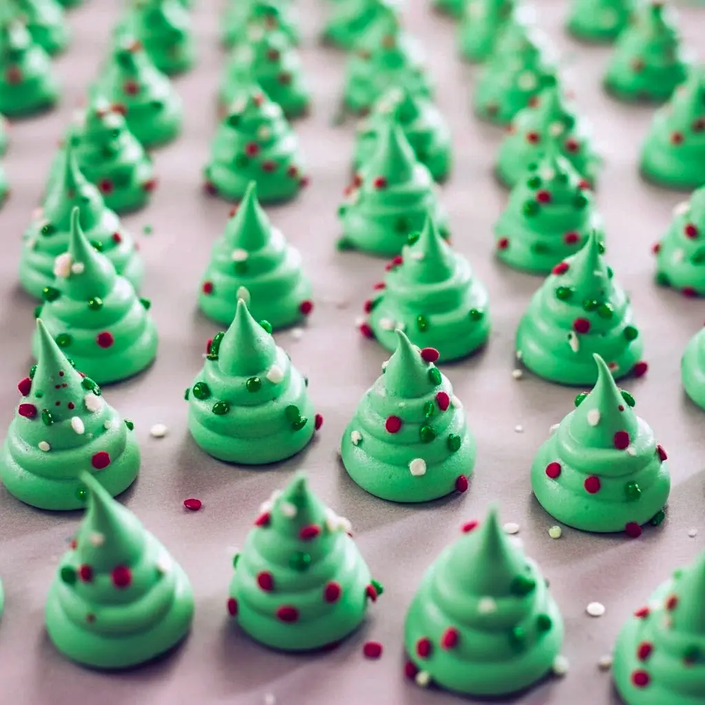 Close-up of piped green meringue Christmas tree cookies on parchment, sprinkled with nonpareils and a few pretzel trunks visible, cooling on a sheet pan, Meringue Christmas Trees.