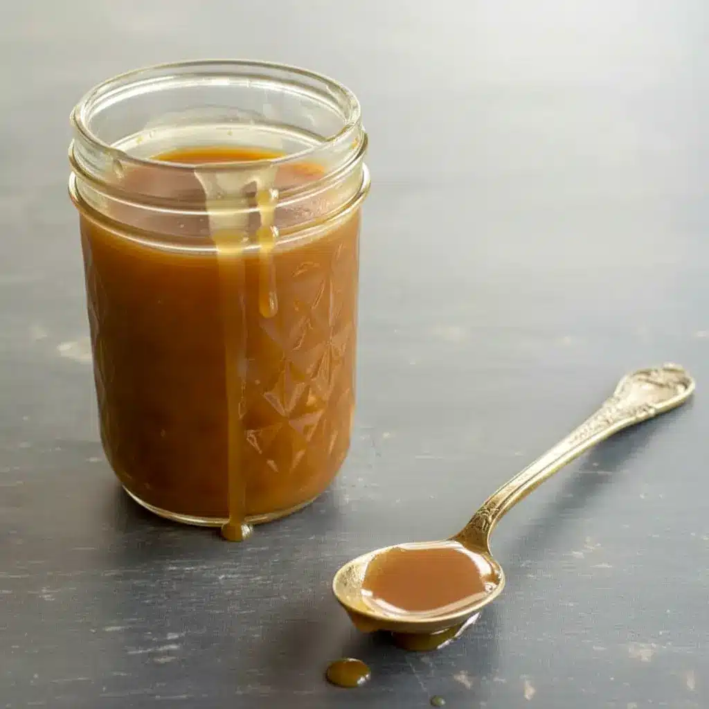 Keto Salted Caramel spoon pouring into a jar, glossy and thick; caption notes Low Carb Keto Caramel Sauce Recipe, nearby are bite-size Keto Caramel Fat Bomb Recipe squares and a dish labeled Keto Candy; the photo sits among jars labeled Keto Sauces and a stack of Breakfast Low Carb pancakes, suggesting easy Keto Dessert Easy and other Low Carb Sweets for Joy Filled Eats.