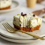 Square no-bake cannoli cheesecake bars on a serving plate, topped with mini chocolate chips and chopped pistachios.