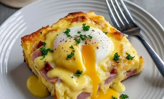 Pan of golden Eggs Benedict Casserole with warm hollandaise, chopped parsley, and visible chunks of English muffin and diced Canadian Bacon.