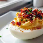 Close-up of halved deviled eggs piped with creamy yolk filling, topped with crumbled bacon, smoked paprika, and chopped chives on a white serving platter, Stuffed Deviled Eggs.