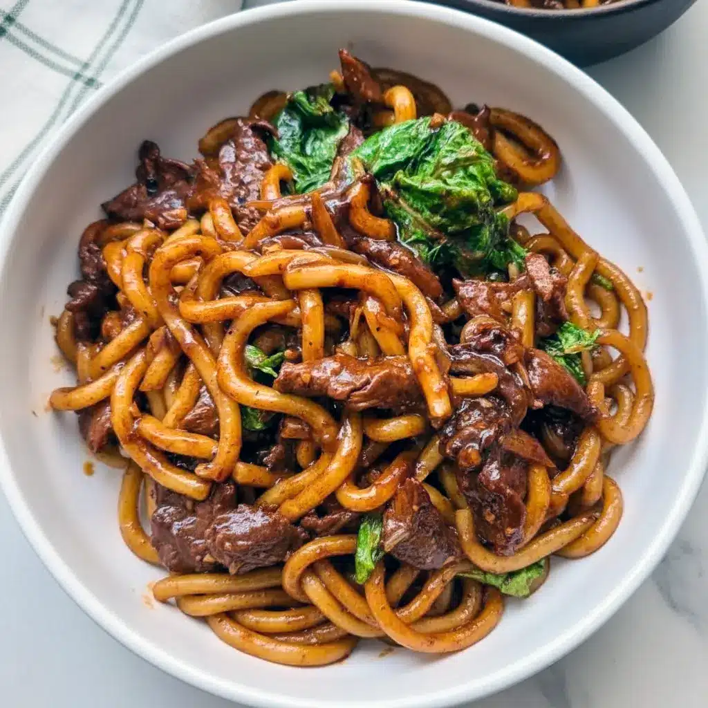 Close-up of black pepper beef udon: glossy noodles with sliced beef, onions, and cabbage coated in a dark pepper sauce, topped with chopped green onions, Asian Recipes.