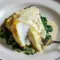 Close-up of Foil-Baked Chilean Sea Bass With Lemon Parmesan Cream Sauce on a white plate: golden, flaky fish over wilted garlic spinach, drizzled with creamy lemon-parmesan sauce and lemon zest.