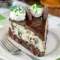 Desserts To Make — close-up of a slice of Thin Mint Cheesecake showing a mint-green cheesecake layer over a chewy brownie crust, glossy chocolate topping, and chopped mint-cookie garnish.