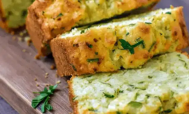 Slice of Zucchini Cheddar Bread on a wooden board, showing gooey cheddar and flecks of green zucchini.