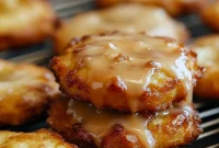 Golden baked apple fritters dotted with apple pieces, drizzled with vanilla glaze and dusted with cinnamon, arranged on a parchment-lined baking sheet.