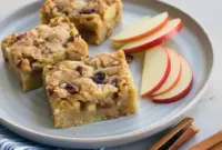 Square apple blondie bars studded with apple pieces and pecans on a wooden board, one piece topped with a scoop of vanilla ice cream.