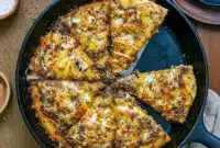 Ultimate Patty Melt Pizza - Pizza With Ground Beef Twist Overhead shot of a cheesy Pizza With Ground Beef topped with caramelized onions and melted cheddar in a cast iron skillet — a cozy Deep Dish Pizza Recipe inspired by a classic patty melt.