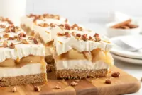 Top view of layered no-bake apple lasagna dessert with graham cracker crust, creamy filling, apple pie topping, and pecans in a glass dish.