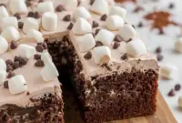 Rich hot cocoa poke cake topped with fluffy whipped chocolate frosting, melted marshmallows, and chocolate chips, served on a festive holiday dessert table.