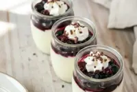 A spoon dipping into a creamy vanilla yogurt protein pudding topped with thawed mixed berries and whipped cream in a small glass container, styled as a high-protein snack or dessert.