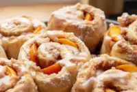 Peach Cobbler Cinnamon Rolls — Sweet, Gooey & Irresistible1 Freshly baked Peach Cobbler Cinnamon Rolls in a baking dish, topped with creamy glaze and filled with juicy peach cinnamon swirls, creating soft and Gooey Peach Cinnamon Rolls perfect for brunch.