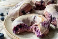 Freshly glazed Blueberry Cake Donuts stacked on a cooling rack with juicy blueberries, perfect for a High Protein Donut Recipe and healthy breakfast treat.